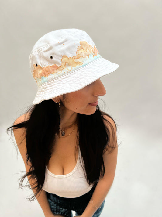 side view of woman wearing white bucket hat painted with cream and light blue colored clouds 