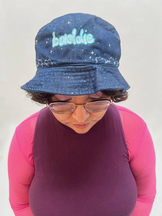 Front angle of woman wearing denim hat painted with word baddie on front 
