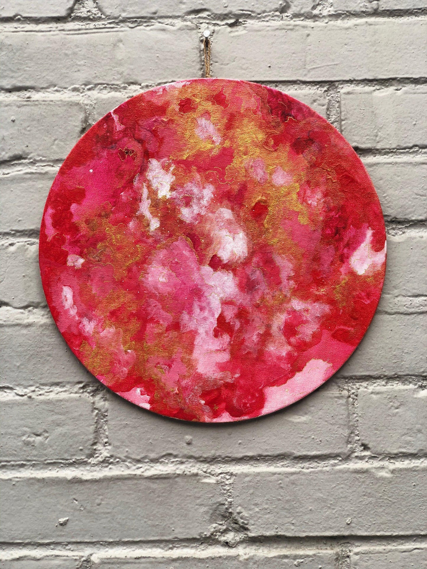 round painting with pink, white, and gold hanging on grey brick wall