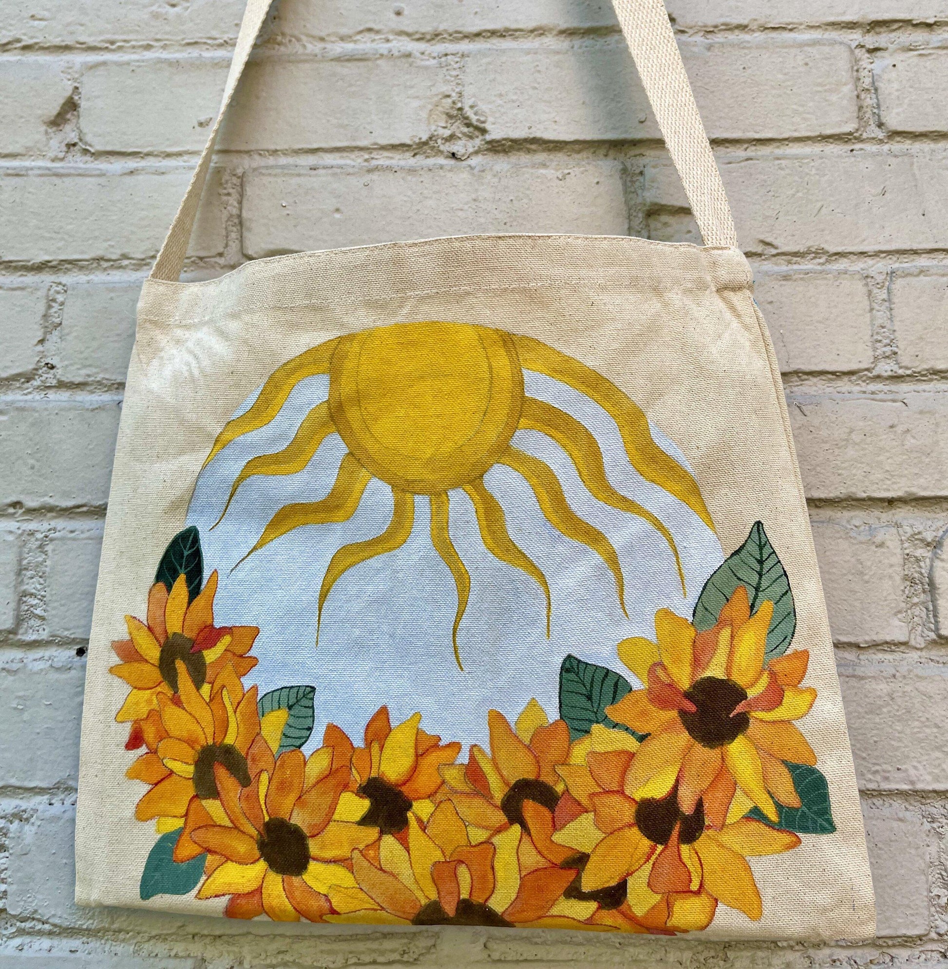 close up of canvas tote with round painting of sunflowers and sun hanging on grey brick wall 