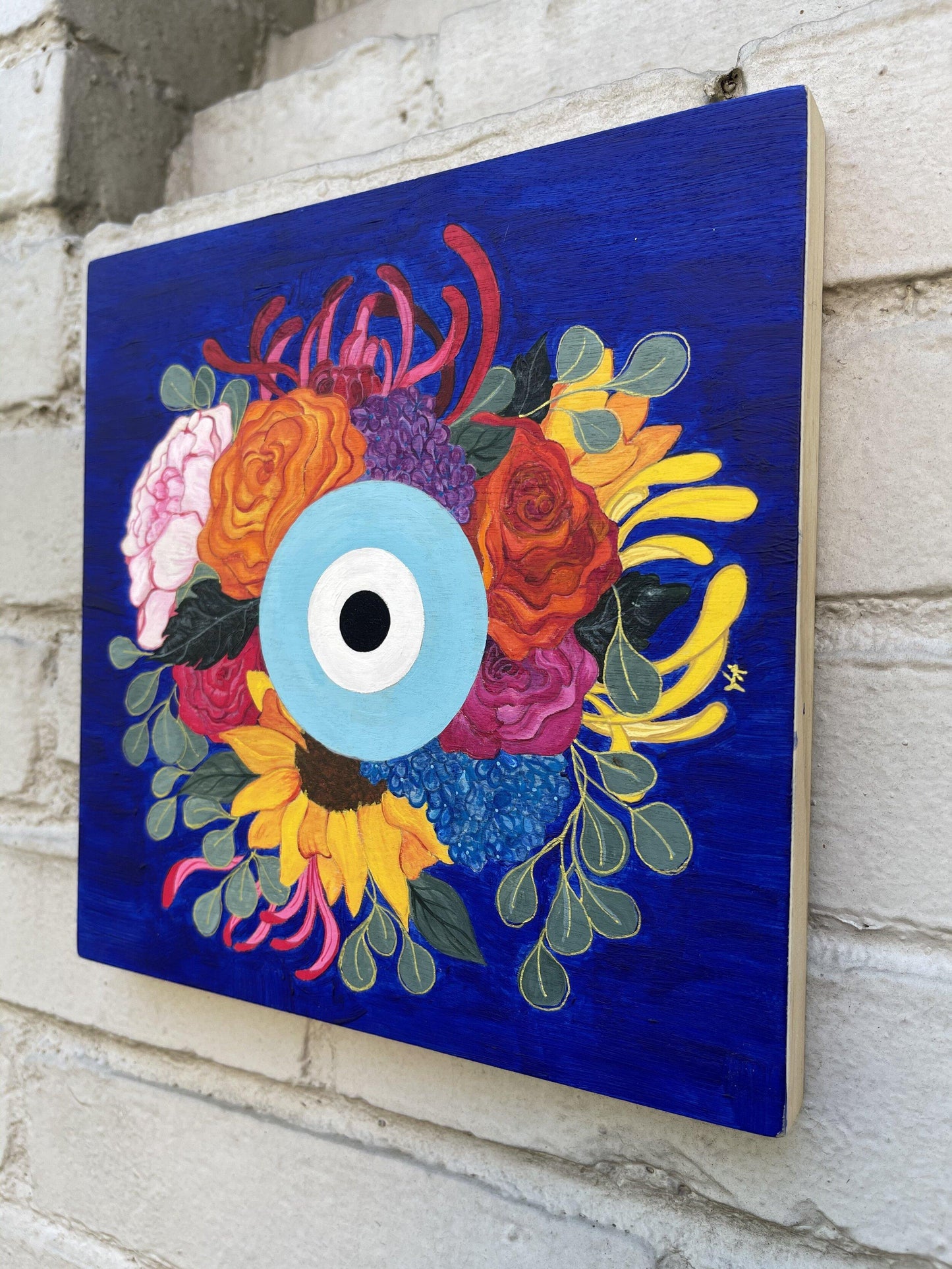 side view painting of round eye surrounded by roses, sunflower, peonies, chrysanthemums, and eucalyptus leaves on grey brick wall