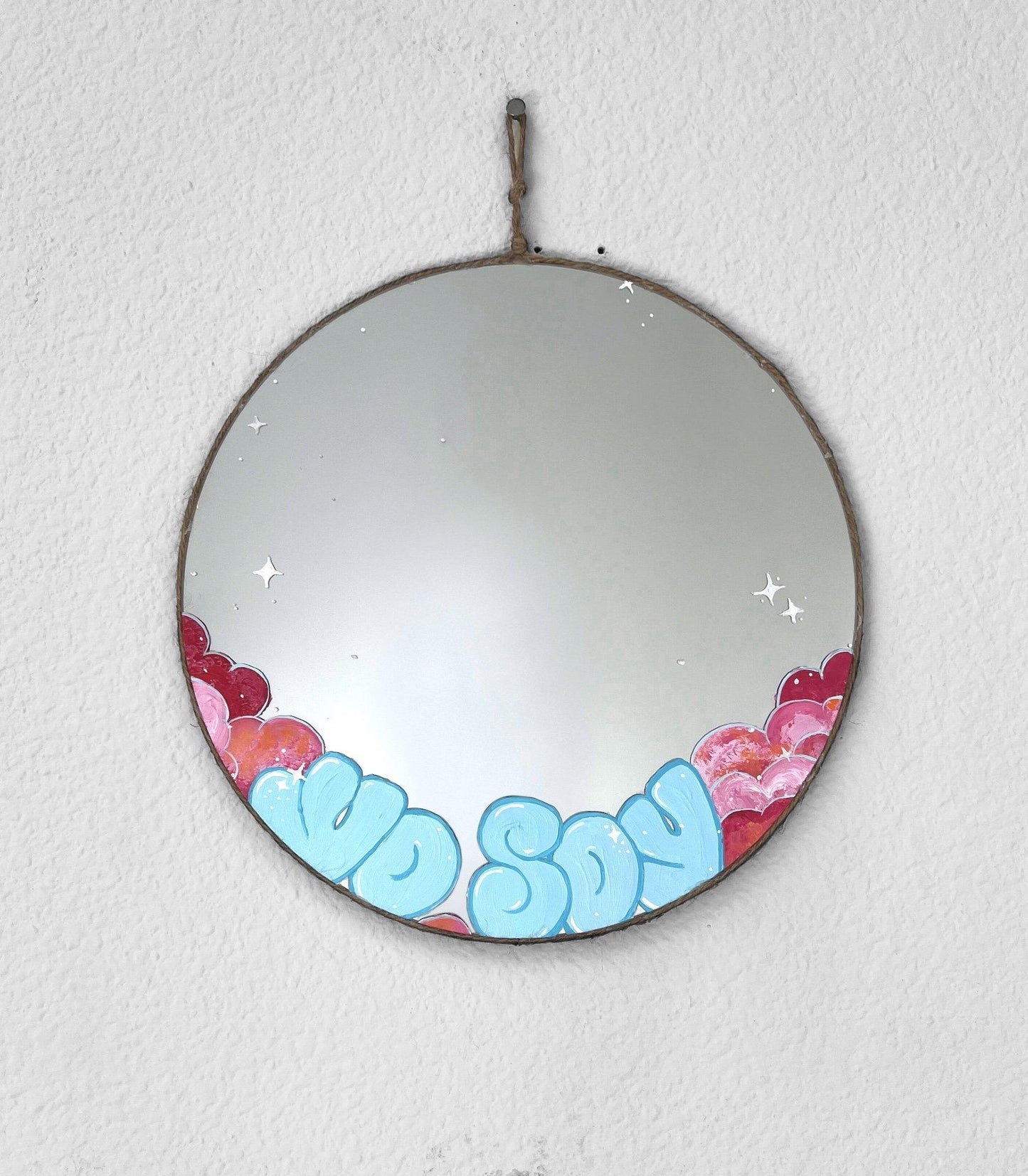 round mirror with light blue writing and pink clouds on bottom hanging on white wall